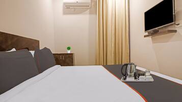 Quarto de hotel com cama, televisão na parede, janela com cortinas e pequena mesa com chaleira e chávenas.