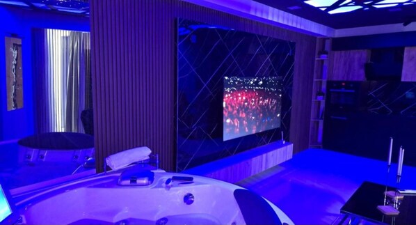 A room with a large screen displaying a crowd, a bed with a white headboard, and a small table with a chair.