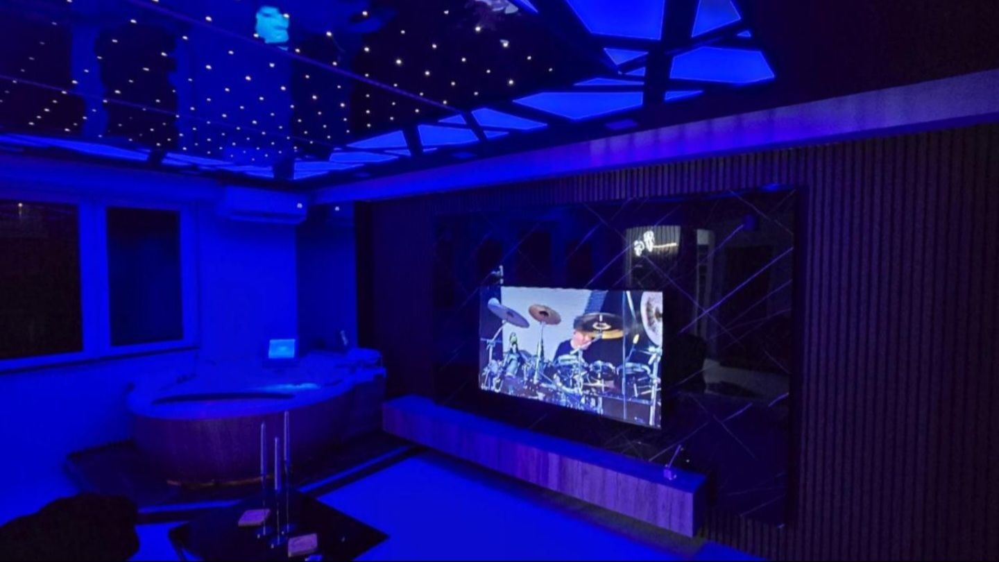 A room with a large screen displaying a band performance, a bar area, and a ceiling with a geometric light fixture.