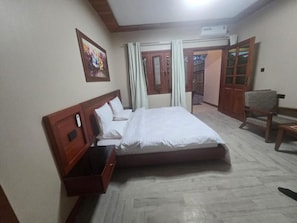 A hotel room with a bed, a chair, a wooden cabinet, a picture on the wall, and a door leading to a balcony. - Across the Nile Resort Jinja (Jinja)