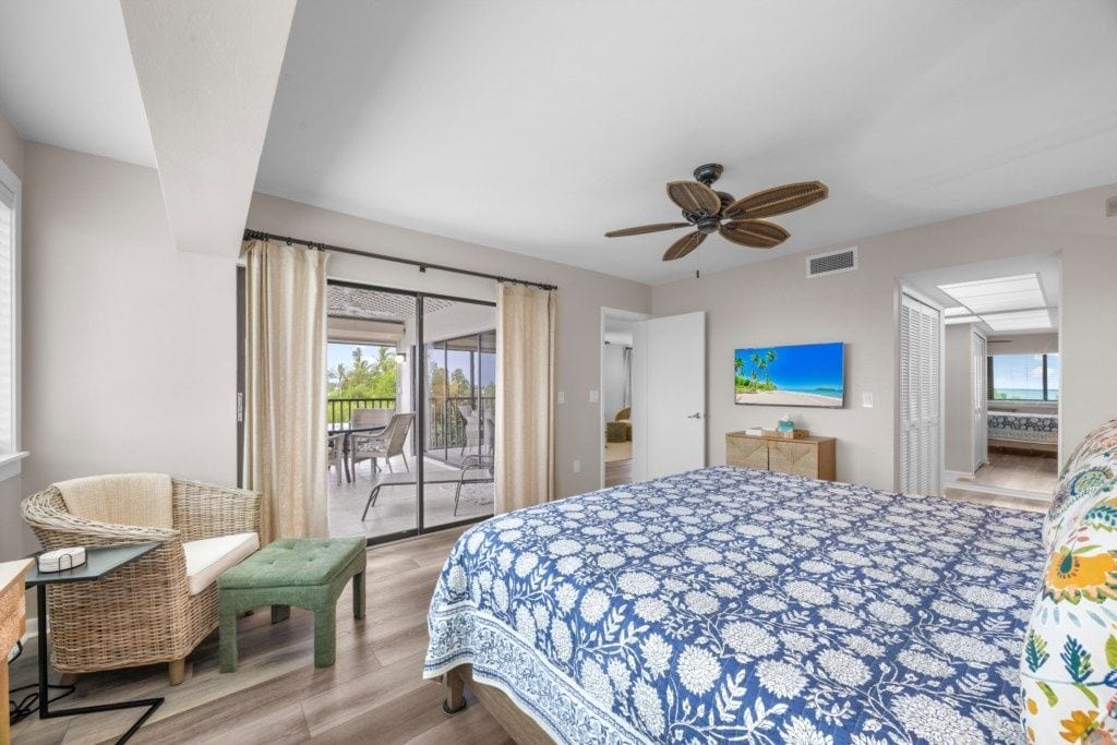 A bedroom with a large bed, a ceiling fan, a television, and a view of the outdoors.