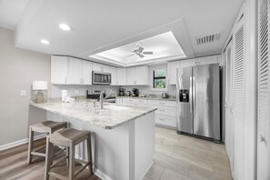 A modern kitchen with white cabinetry, a central island, stainless steel appliances, and a ceiling fan. - Sunset Captiva Bayside Unit 104-new 2 Bedroom Condo (Captiva)