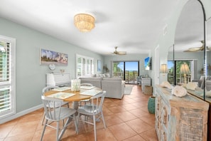 A spacious living area with a dining table, chairs, and a sofa. There is a large window with blinds, a ceiling fan, and a decorative mirror. - Captiva Shores 6a 2 Bedroom Condo (Captiva)