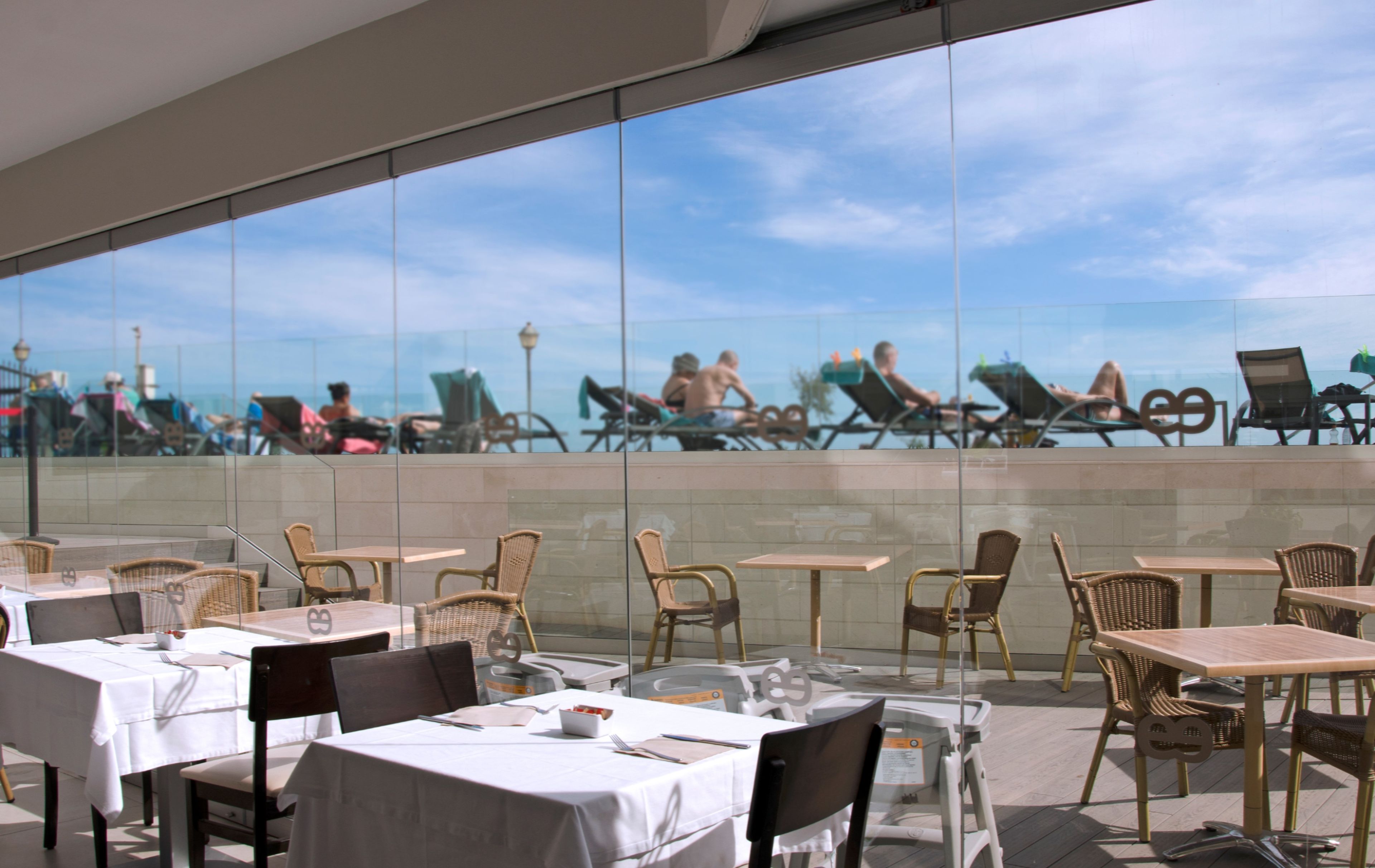 Un espace de restauration en plein air, avec des tables et des chaises, donnant sur une piscine où des personnes prennent des bains de soleil.