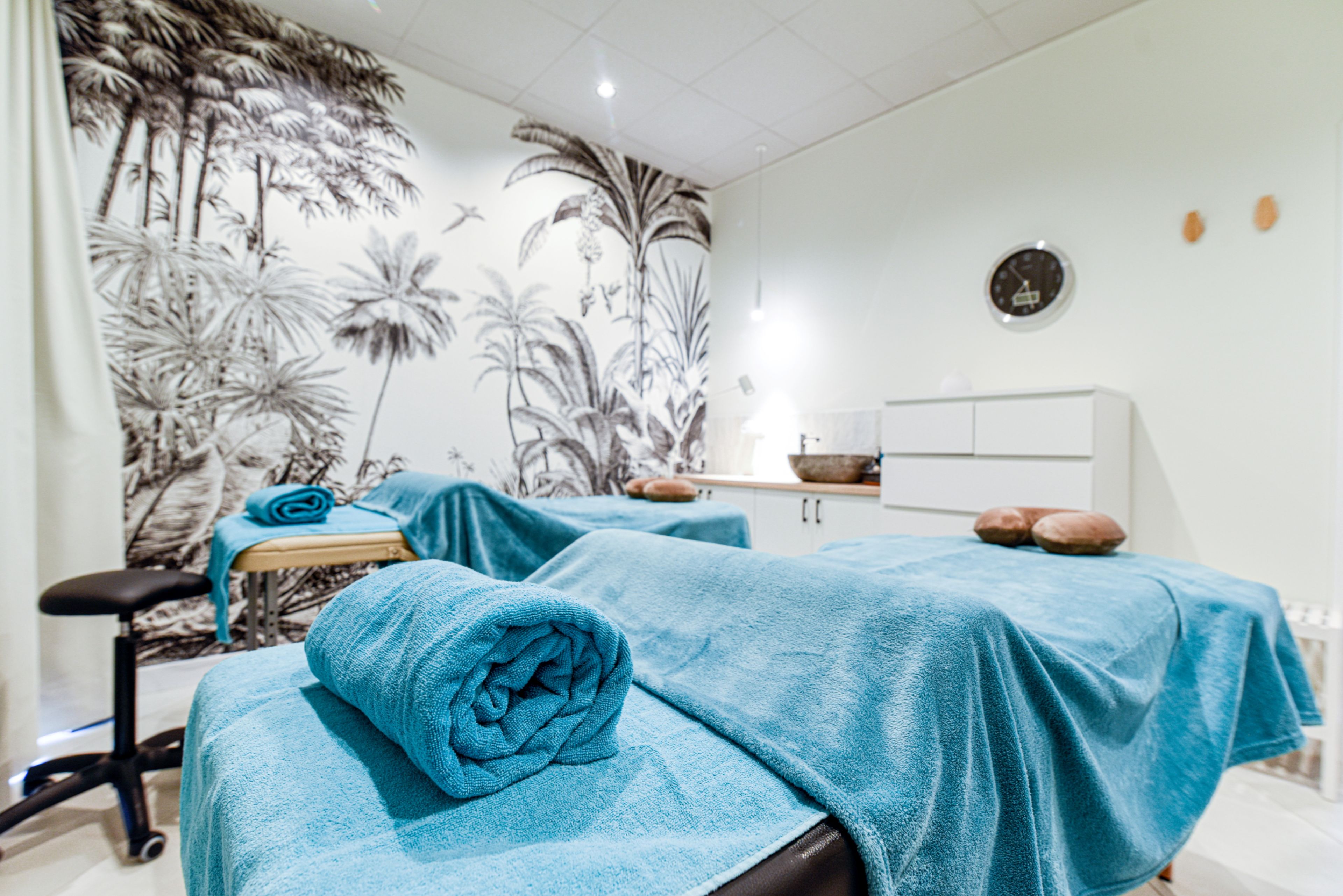 A spa room with two massage tables covered in blue towels, a massage chair, a wall mural of palm trees, a clock, and a small shelf with decorative items.