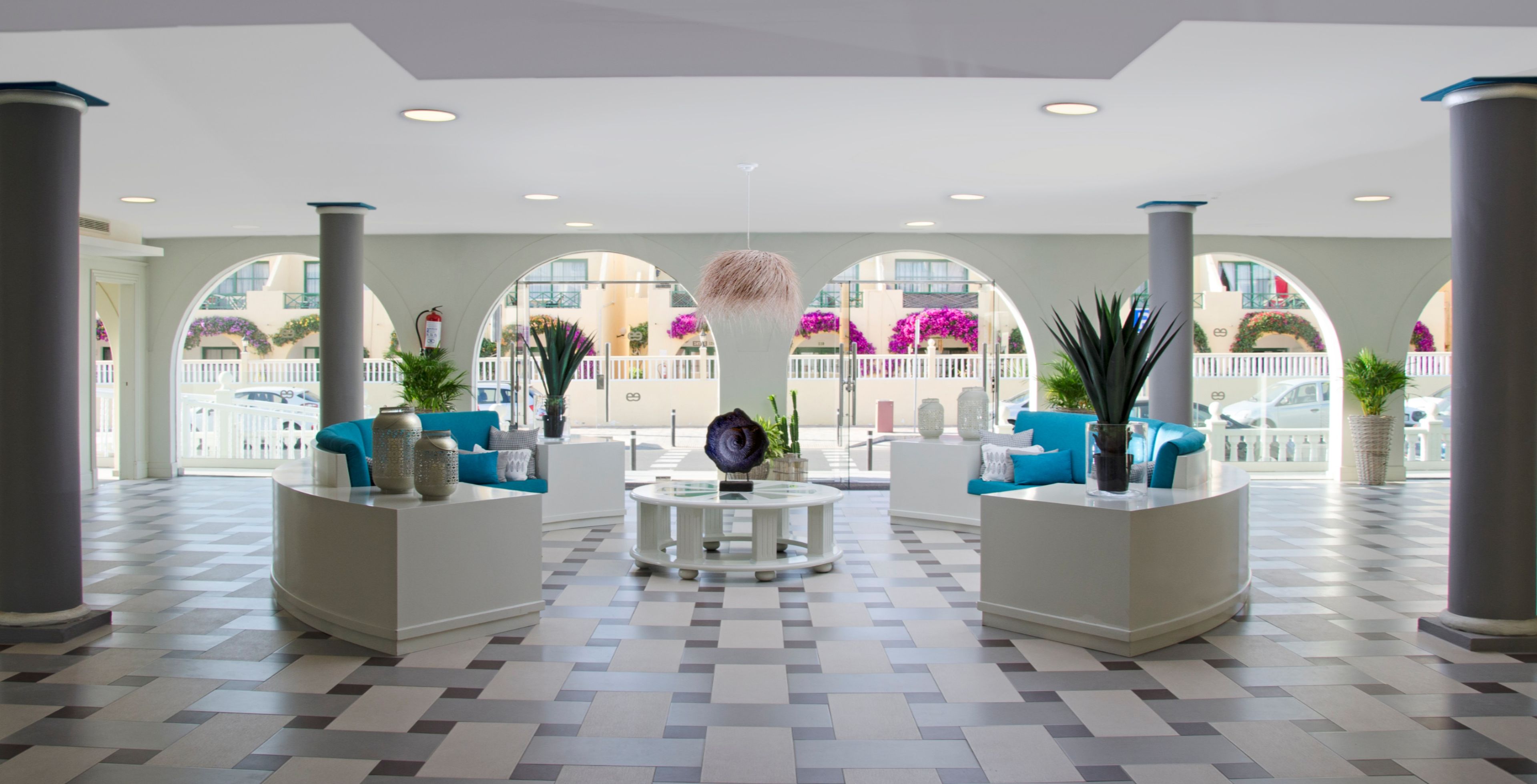 A hotel lobby with a checkered floor, two seating areas with blue sofas, a central coffee table, and large windows with arched tops.