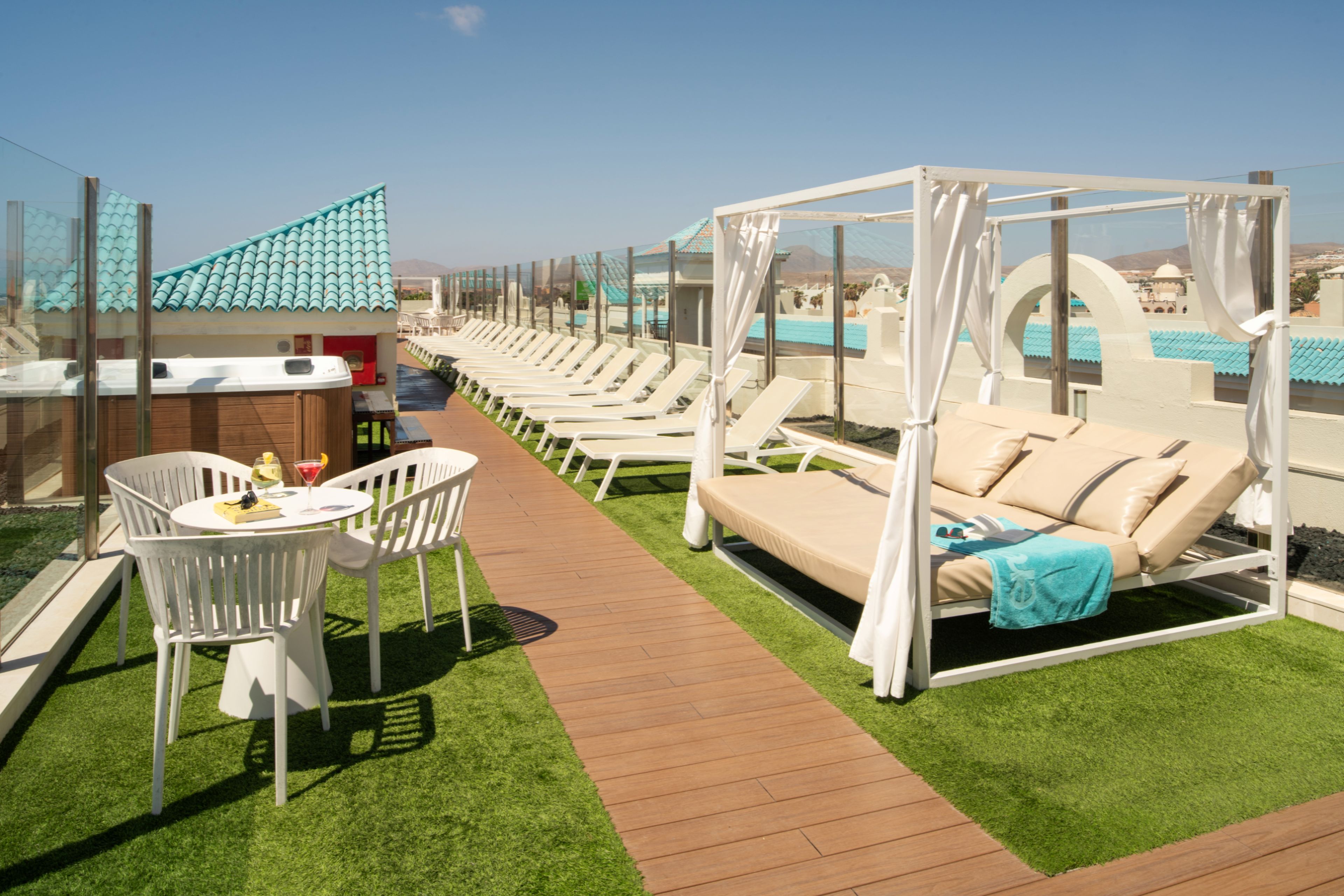 A rooftop lounge area with a pool, white lounge chairs, a white table with drinks, and a canopy bed.