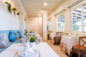 Un comedor con mesas preparadas para el servicio, un sofá con cojines estampados y vistas a través de amplios ventanales.
