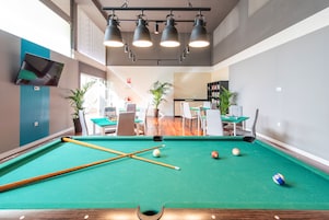 Una mesa de billar con bolas y tacos, un área de estar con mesas y sillas, y una estantería en una habitación de diseño moderno.