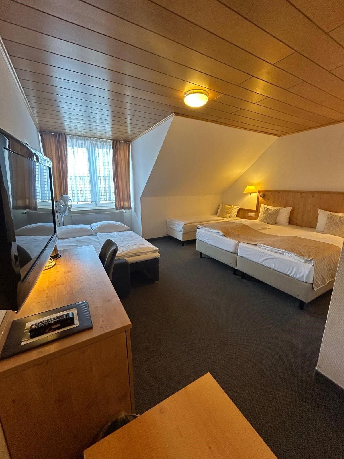 Ein Hotelzimmer mit Holzbalkendecke, einem Schreibtisch mit Fernseher, zwei Betten und einem Fenster mit Vorhängen.