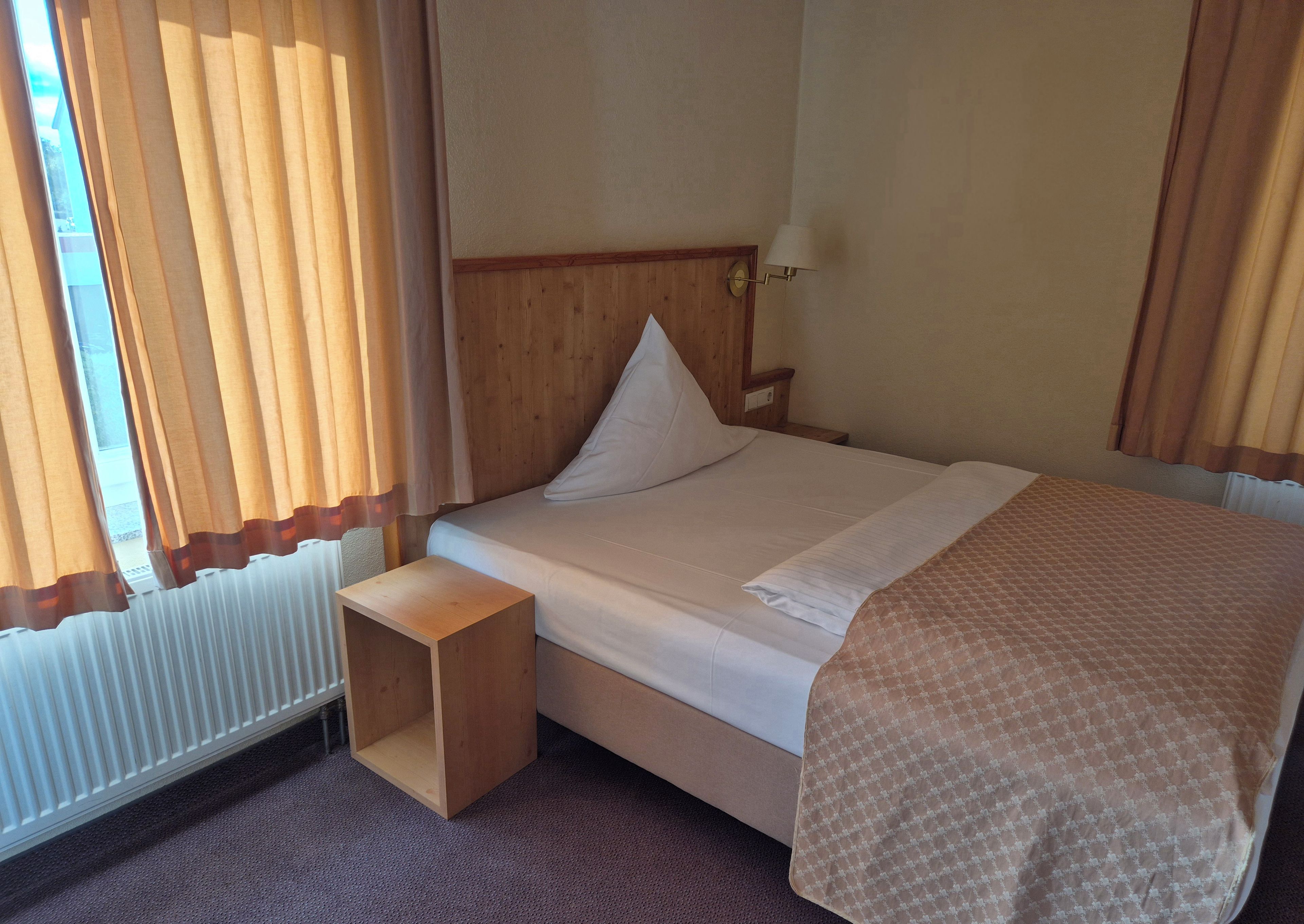 Ein Hotelzimmer mit Bett, Nachttisch, Lampe und einem Fenster mit Vorhängen.