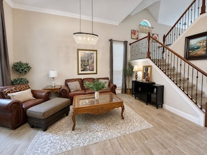 A spacious living room with a brown leather sofa, a glass coffee table, and a staircase with a wooden railing. - 30 Day Rental | Spacious Home with Private Pool | Near Grapevine Lake (Flower Mound)