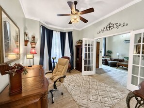 Une pièce dotée d’un ventilateur de plafond, d’un bureau en bois, d’un fauteuil beige, d’un tapis à motifs et d’une vue sur une autre pièce, accessible par une porte ouverte.