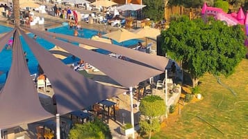 Zona de piscina con múltiples tumbonas y sombrillas, rodeada de vegetación y una estructura que recuerda a un castillo.