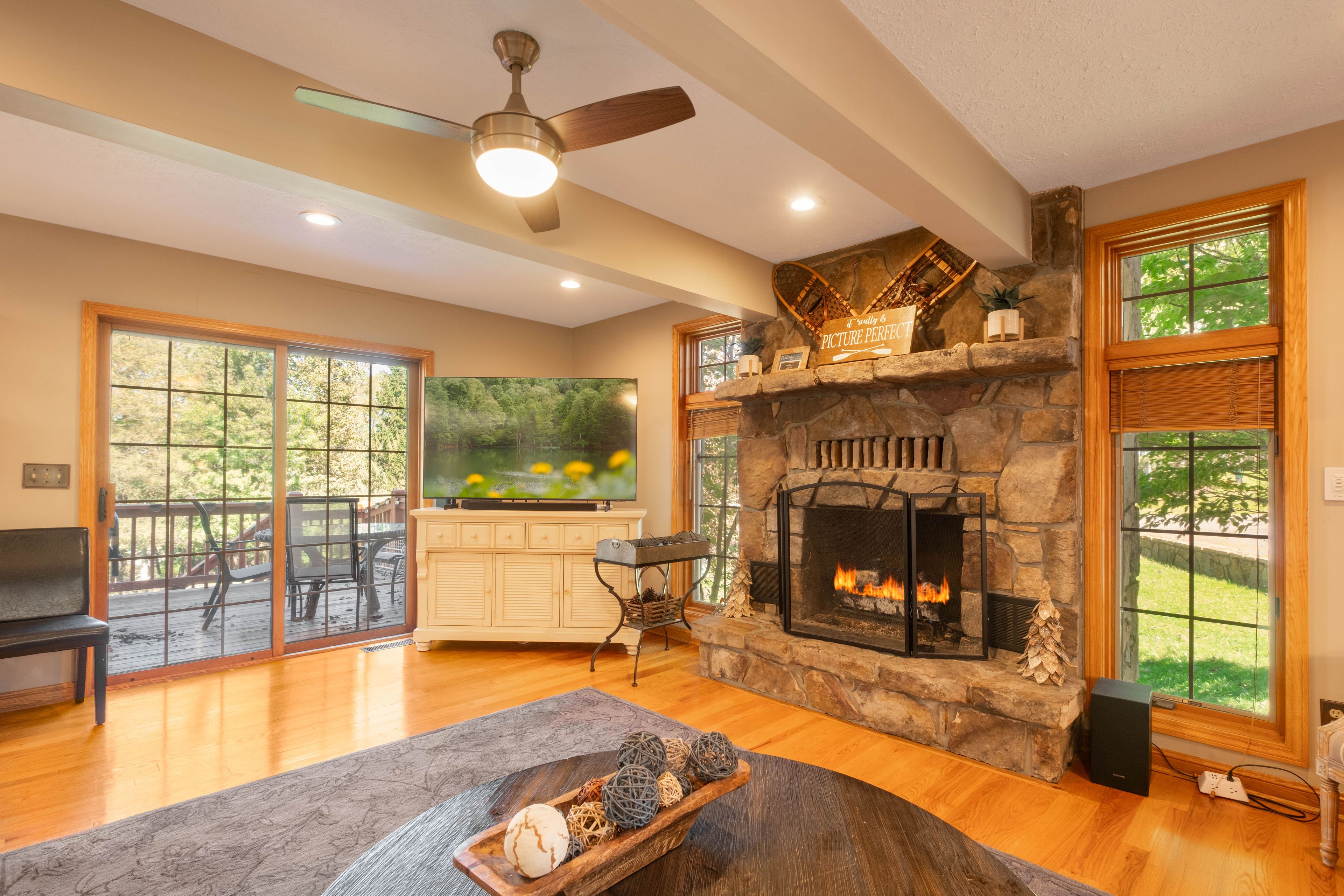 A spacious living room with a stone fireplace, a ceiling fan, and a large window overlooking a deck.