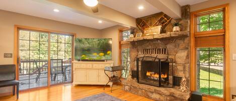 A spacious living room with a stone fireplace, a ceiling fan, and a large window overlooking a deck.
