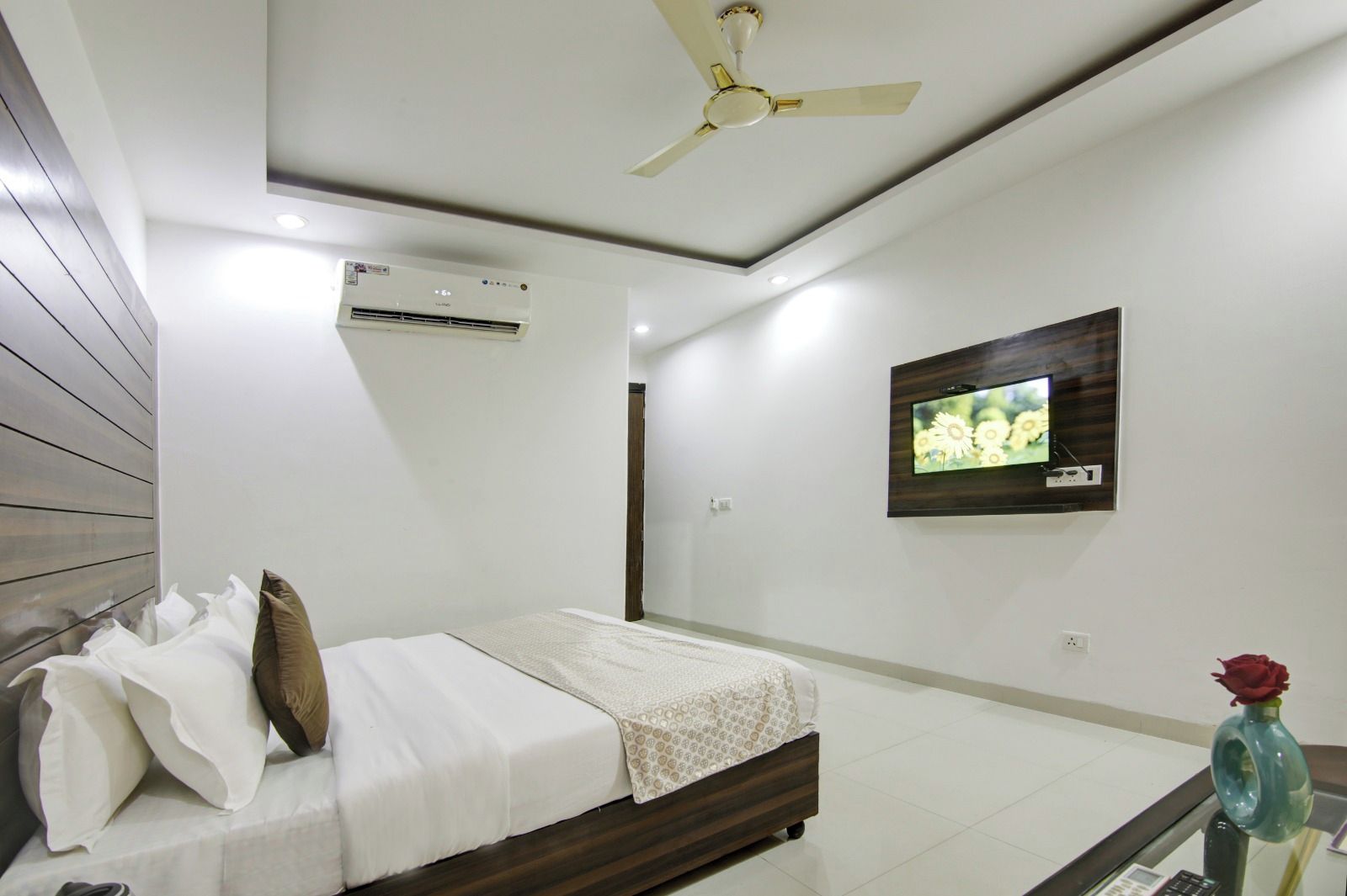 Une chambre à coucher comprenant un lit, un ventilateur de plafond, un appareil de climatisation, un téléviseur fixé au mur et un vase avec une fleur.