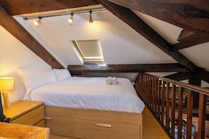 Ein Schlafzimmer mit schrägverdeckter Decke, einem Bett mit weißer Bettwäsche, einem Nachttisch, einer Lampe und einem Fenster mit Jalousien.