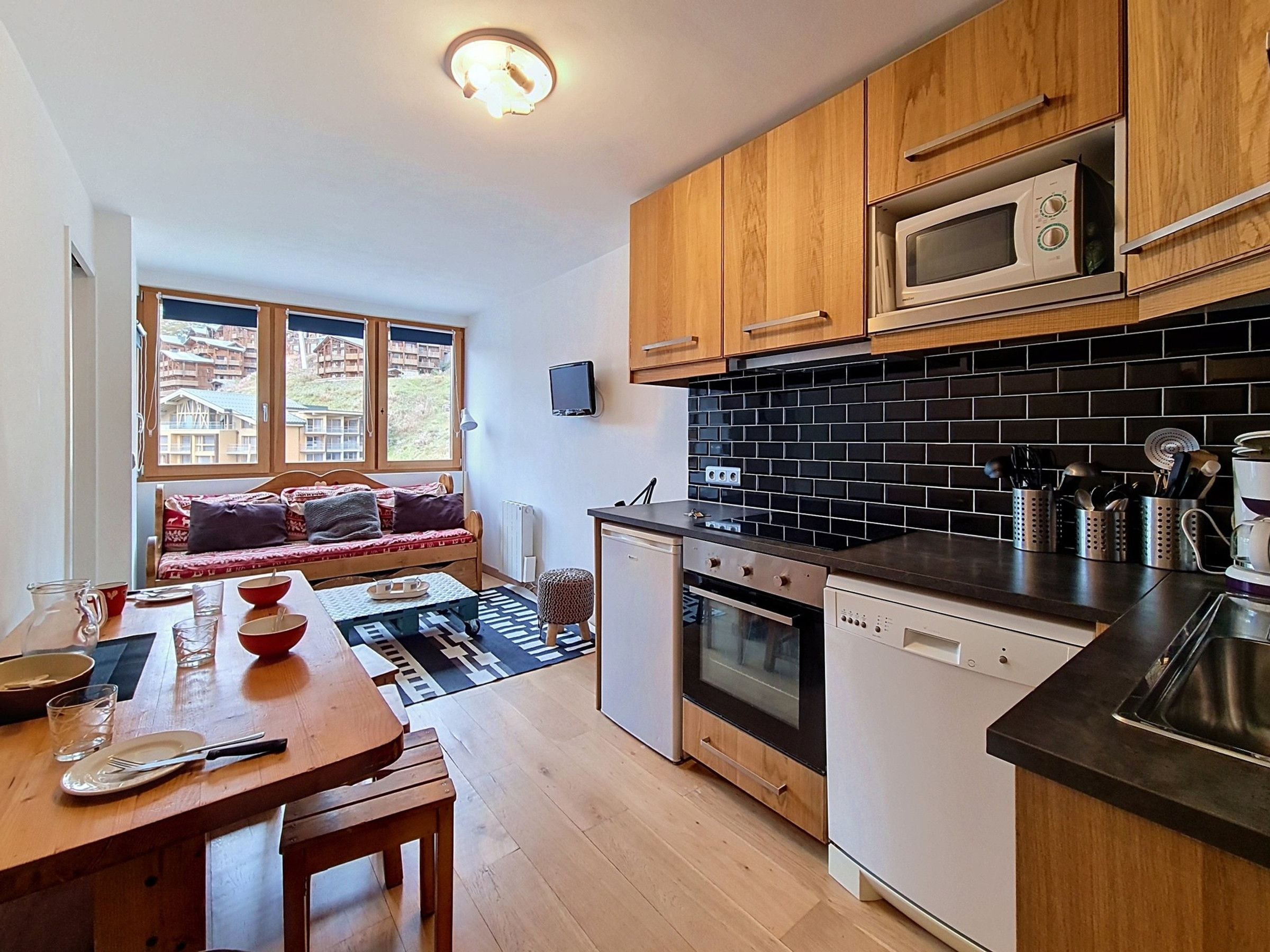 A compact kitchen with wooden cabinets, a microwave, a stove, and a dishwasher. There's a dining area with a table and chairs, and a window with a view of buildings outside.