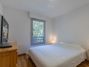 A bedroom with a bed, a desk with a computer monitor, a window with a view of trees, and a nightstand with a lamp. - Appartement spacieux · Balcons · Accès aux pistes par navette (La Salle-les-Alpes)