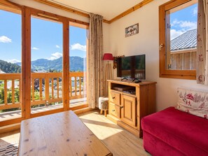 Een kamer met een houten tafel, een rode bank, een tv op een houten dressoir en uitzicht op bergen door grote glazen deuren.