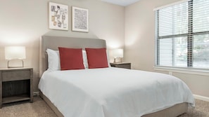 Una cama bien hecha con almohadas blancas y rojas, dos mesitas de noche con lámparas, y una obra de arte en la pared.