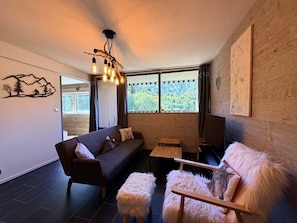 Ein modernes Wohnzimmer mit einer Couch, einem Couchtisch und einem kuscheligen Sessel. Ein Fenster bietet Blick auf grüne Umgebung.
