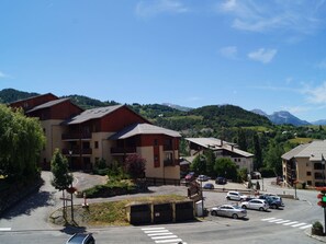 A multi-story building with balconies, a parking area, and a view of green hills and mountains in the background. - Studio en coeur de station (Enchastrayes)
