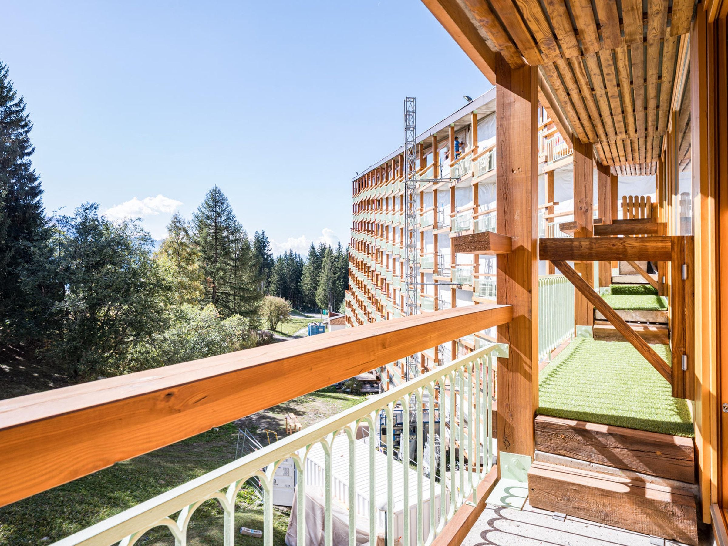 A wooden balcony with a view of a forested area and a building with multiple balconies.