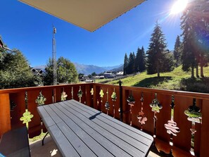 En träveranda med ett bord och stolar, med utsikt över ett bergigt landskap med träd och en klar himmel.