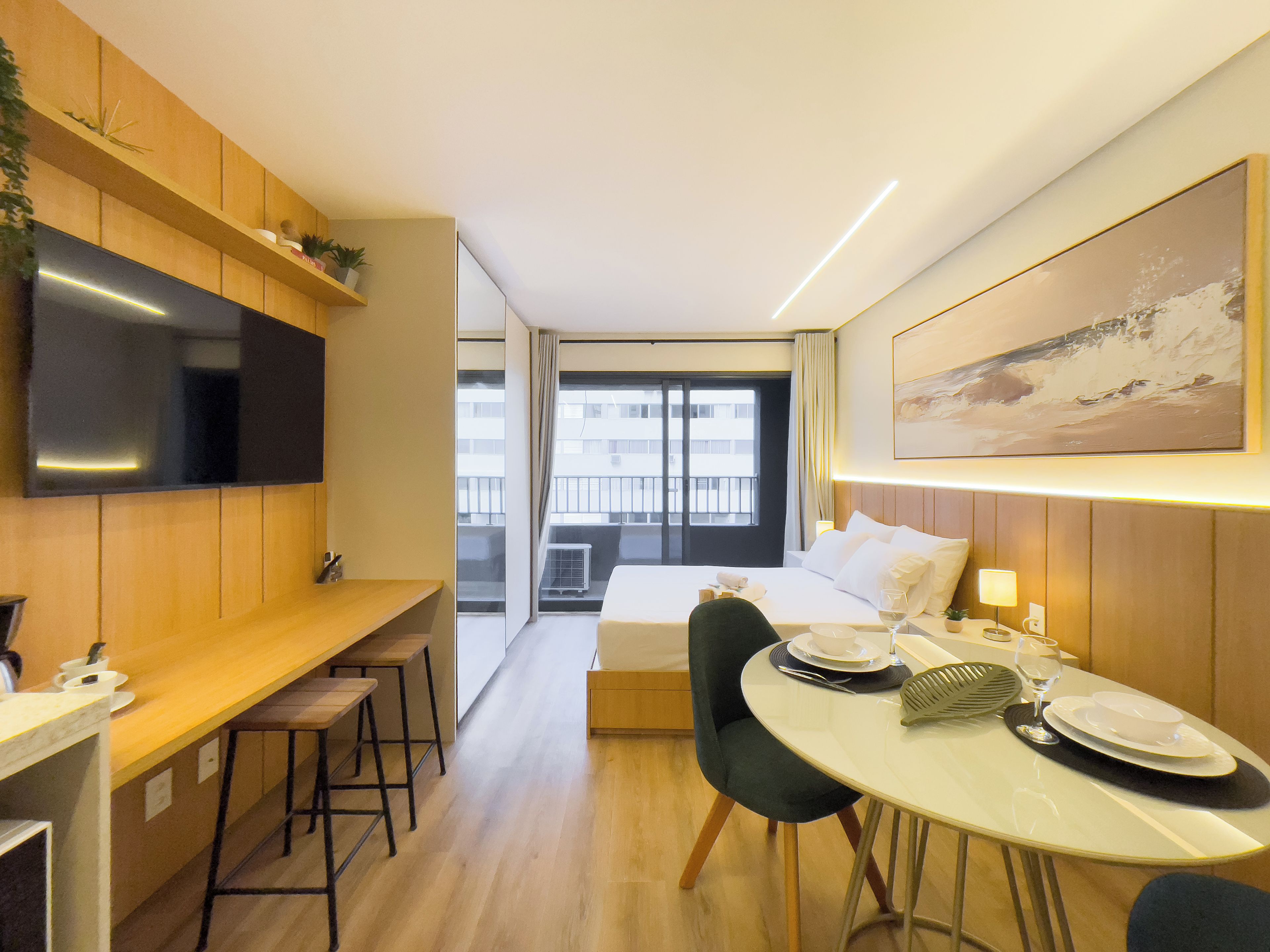 A hotel room with a bed, a dining table with chairs, a TV mounted on the wall, and a window with a view of the city.