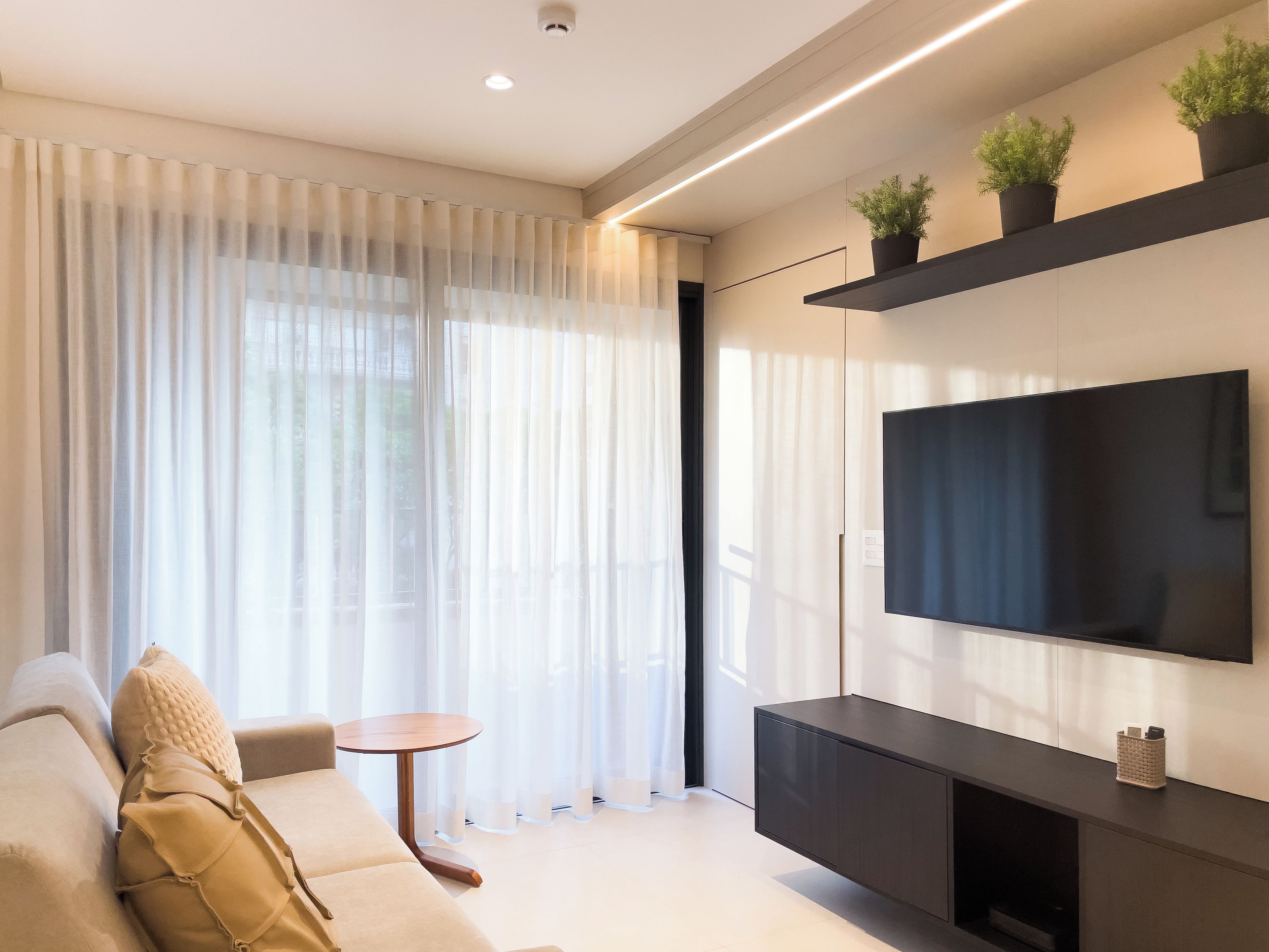 A modern living room with a sofa, a small round table, a flat-screen TV mounted on a wall, and a potted plant on a shelf.