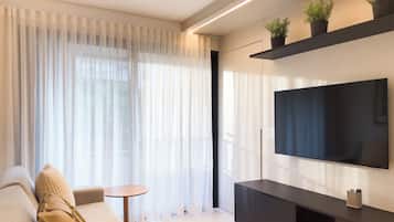 A modern living room with a sofa, a small round table, a flat-screen TV mounted on a wall, and a potted plant on a shelf.