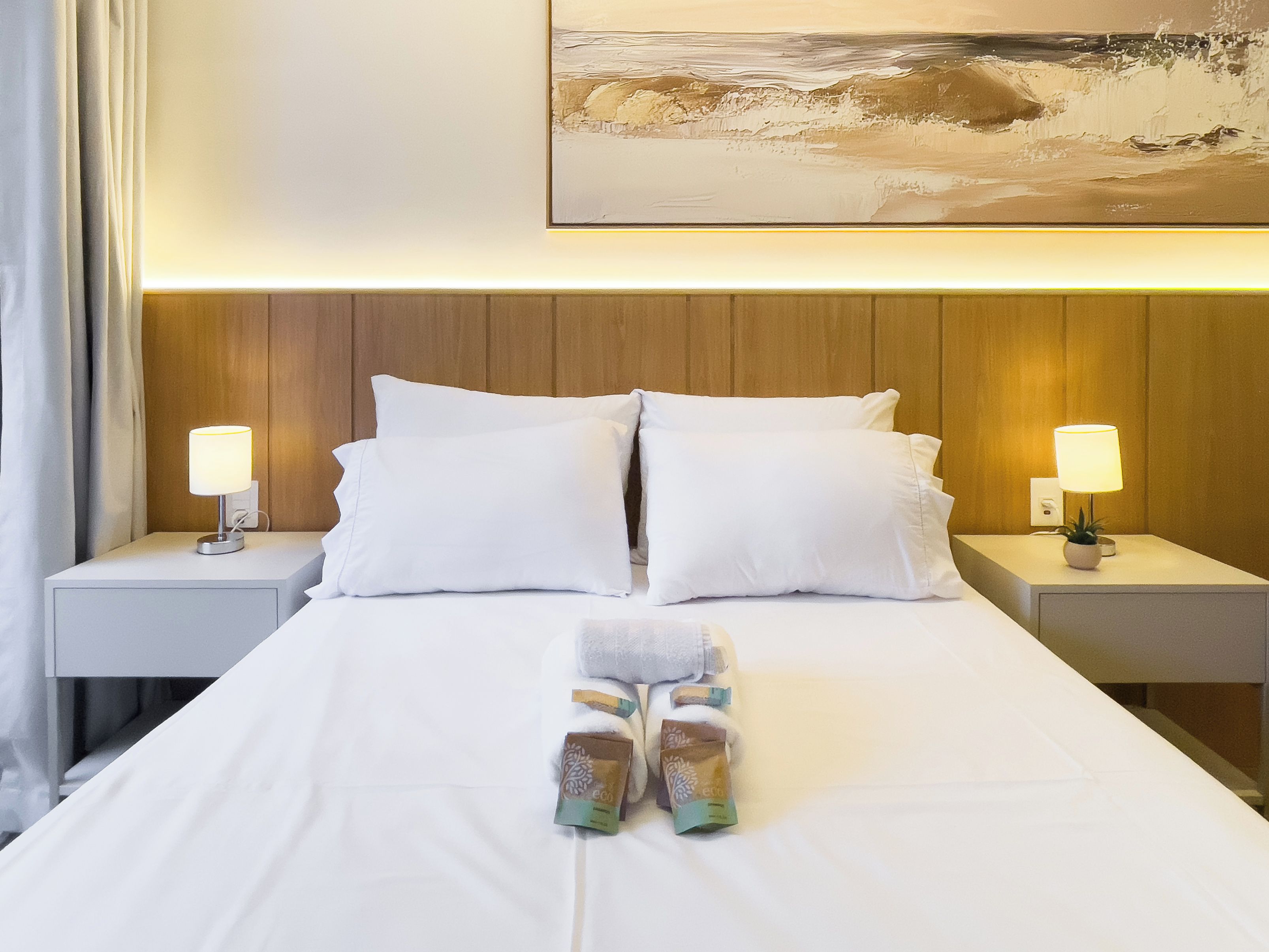 A neatly made bed with white linens, two pillows, and a pair of folded towels. There are two bedside lamps on either side of the bed, and a painting above the headboard.