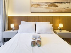 Cama bem arrumada com roupa de cama branca, dois travesseiros e um par de toalhas dobradas. Há duas luminárias de cabeceira de cada lado da cama, e um quadro acima da cabeceira.