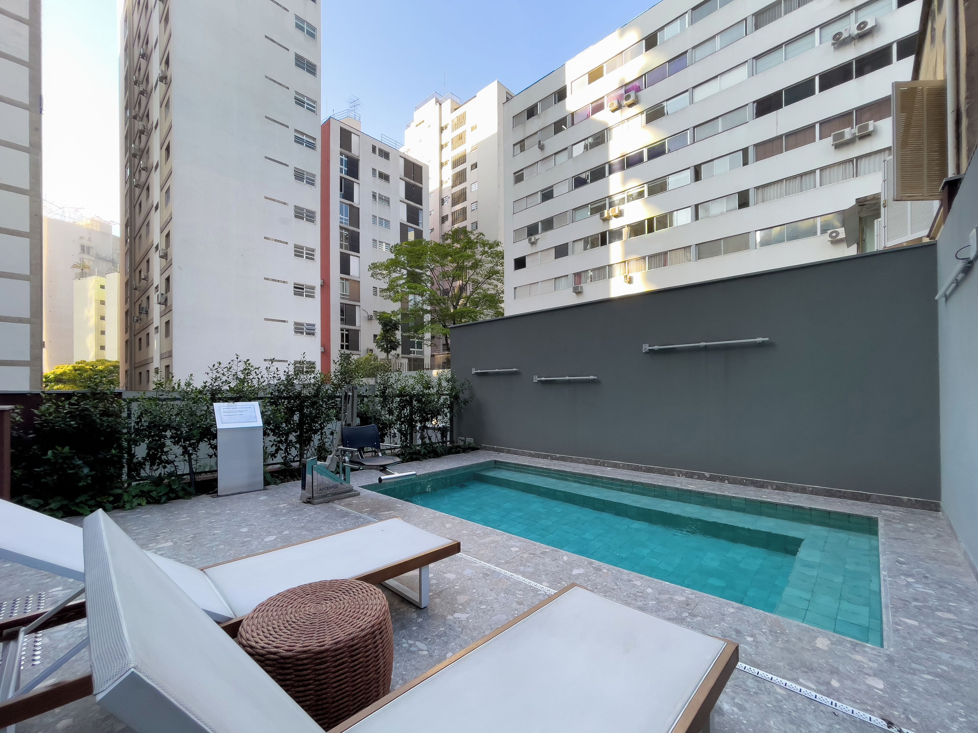 Un área de piscina en la azotea con sillones de descanso y una pequeña mesa, rodeada de edificios altos.