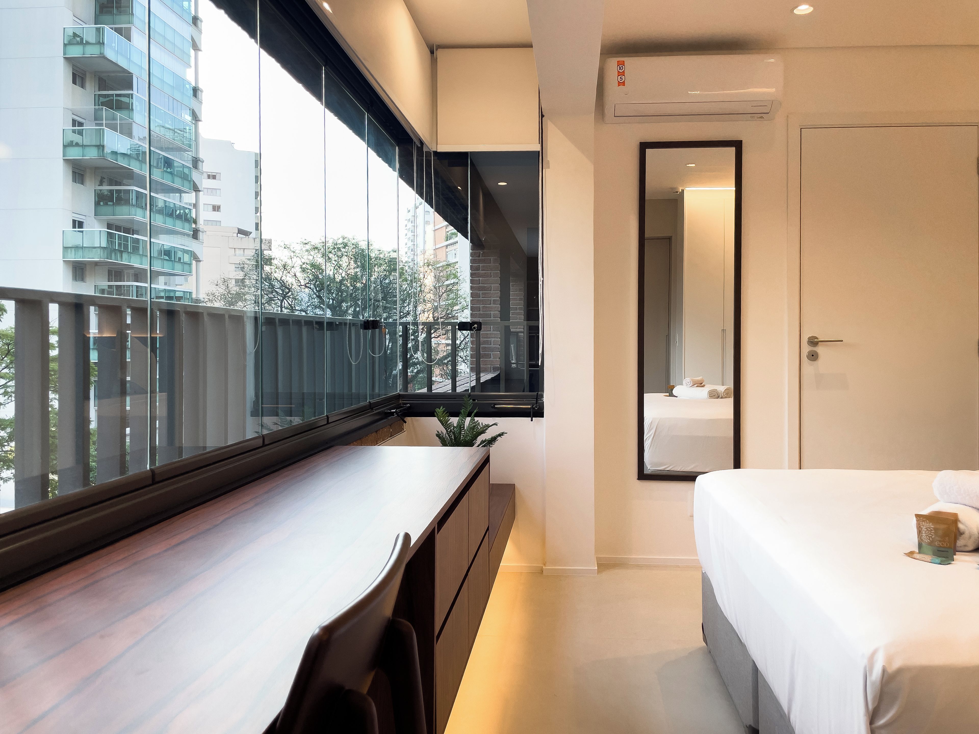 A modern hotel room with a balcony, a bed, a desk, and a large window offering a city view.