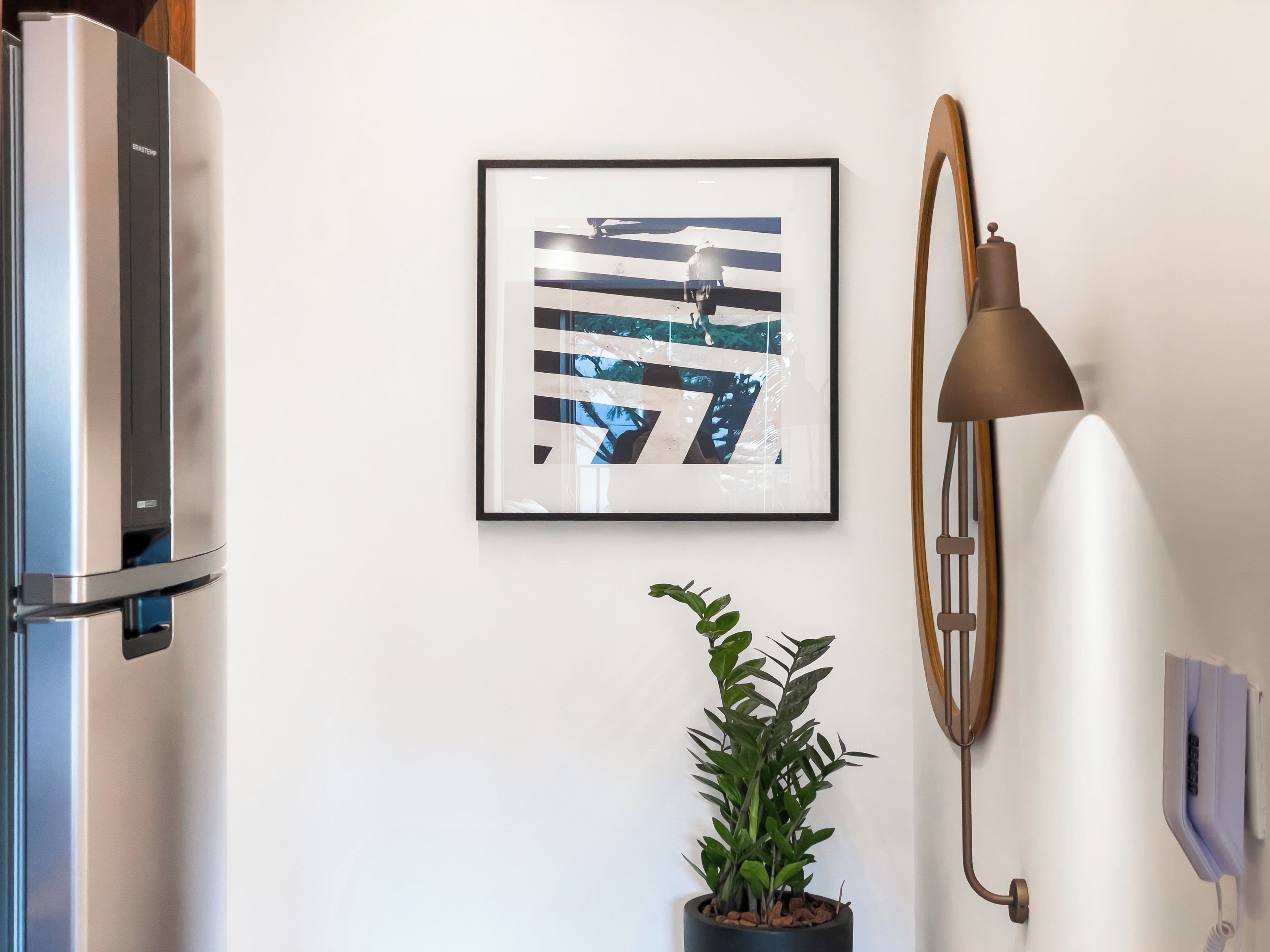 A modern kitchen with a stainless steel refrigerator, a framed abstract painting, a potted plant, and a wall-mounted lamp.