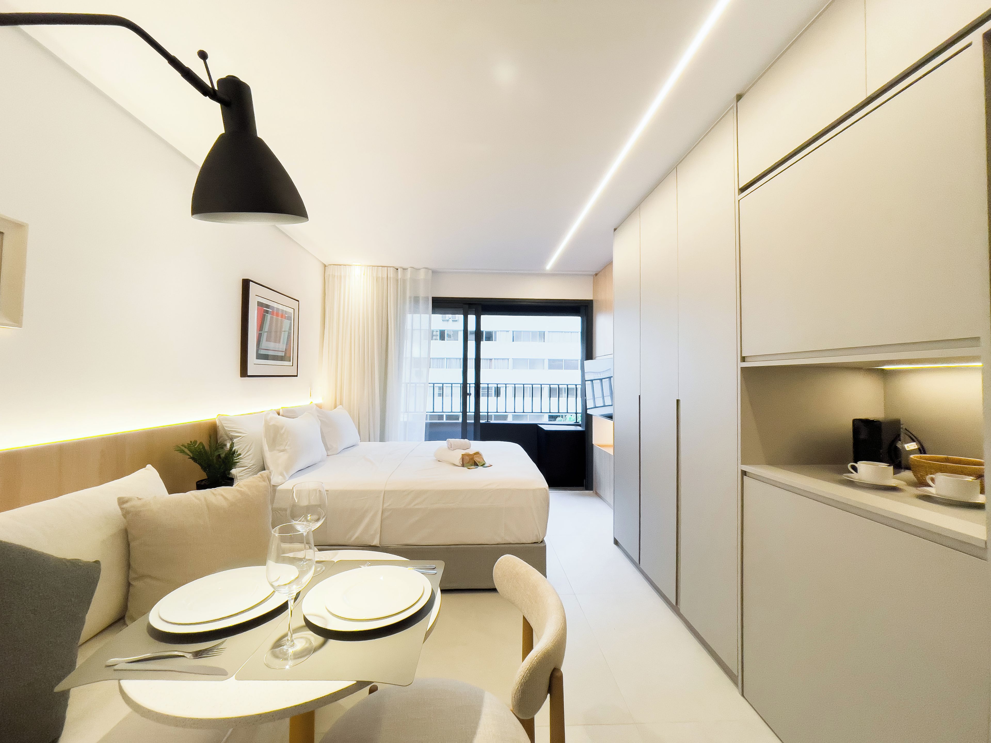 A compact hotel room with a bed, a dining area with a table set for two, a kitchenette, and a balcony with a view.