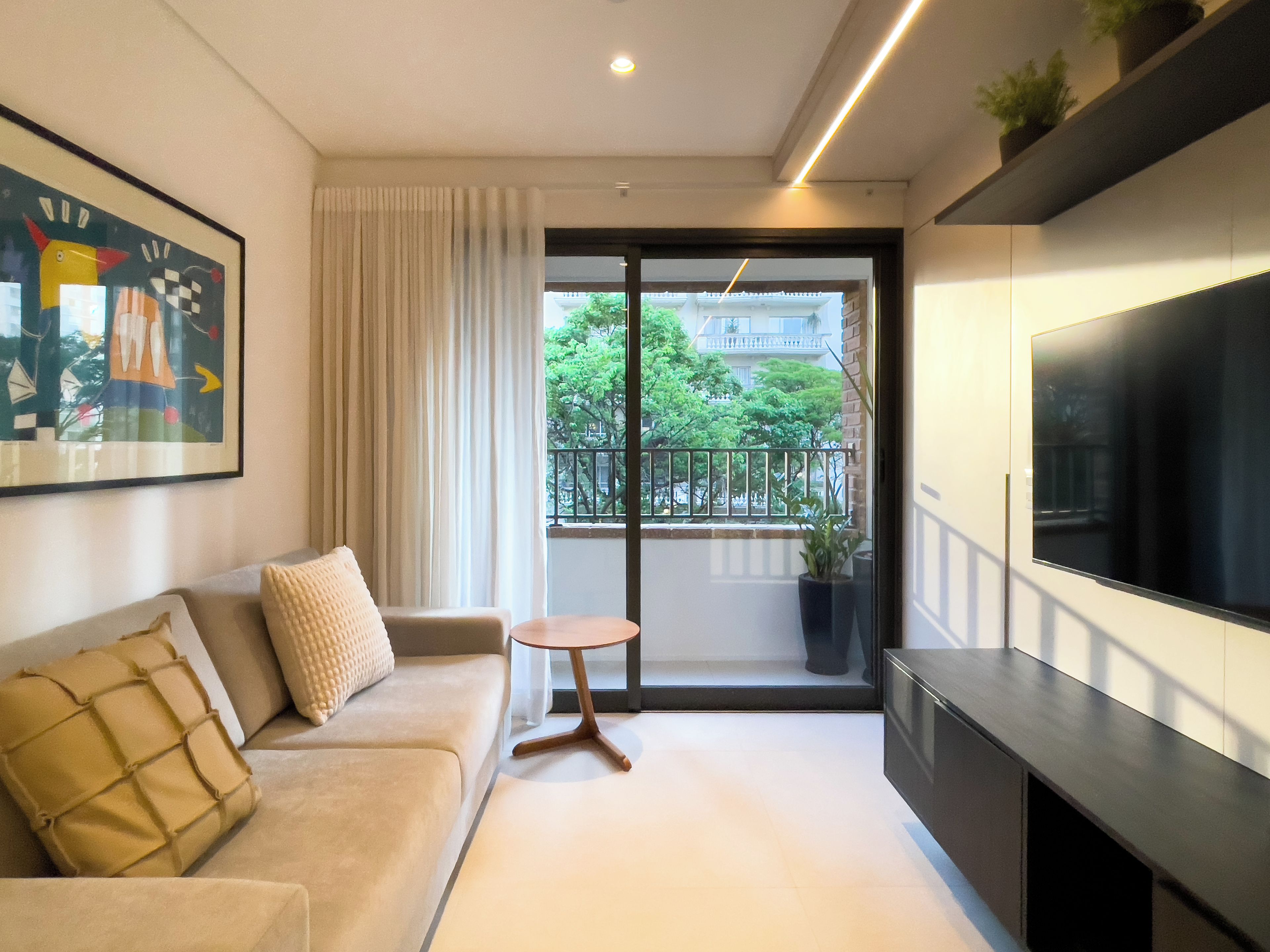 A modern living room with a sofa, a small round table, a TV, and a balcony with a view of greenery and buildings.