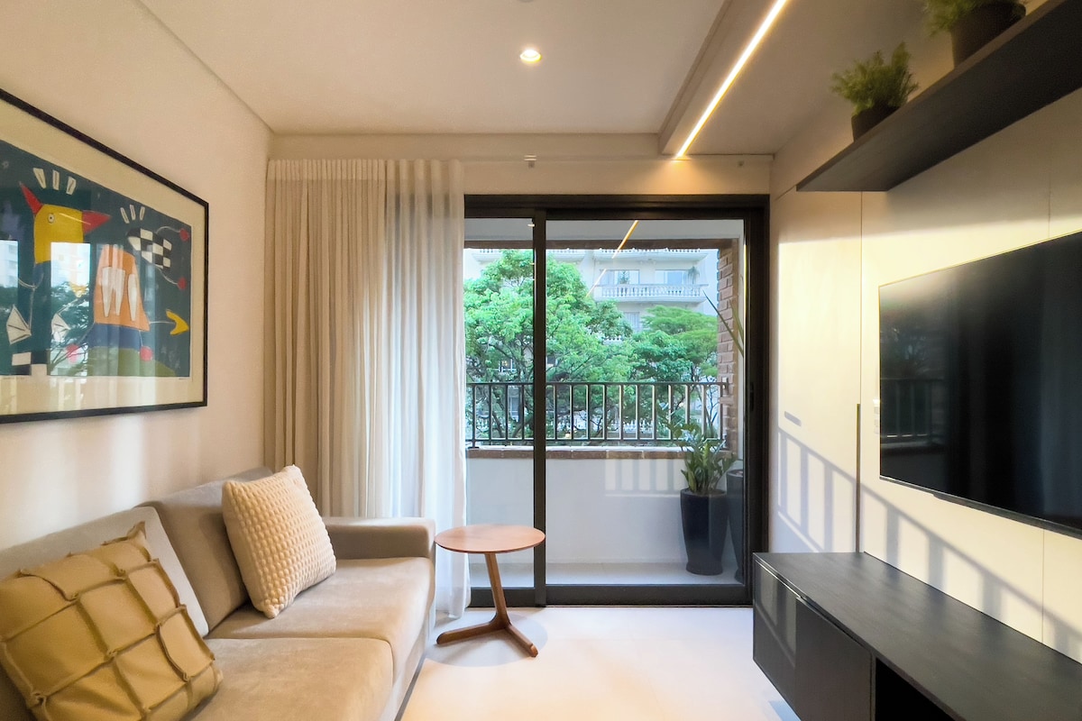 Un soggiorno moderno con divano, un tavolino rotondo, televisione e un balcone vista su verde e edifici.