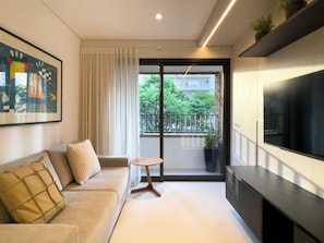 Un soggiorno moderno con divano, un tavolino rotondo, televisione e un balcone vista su verde e edifici.