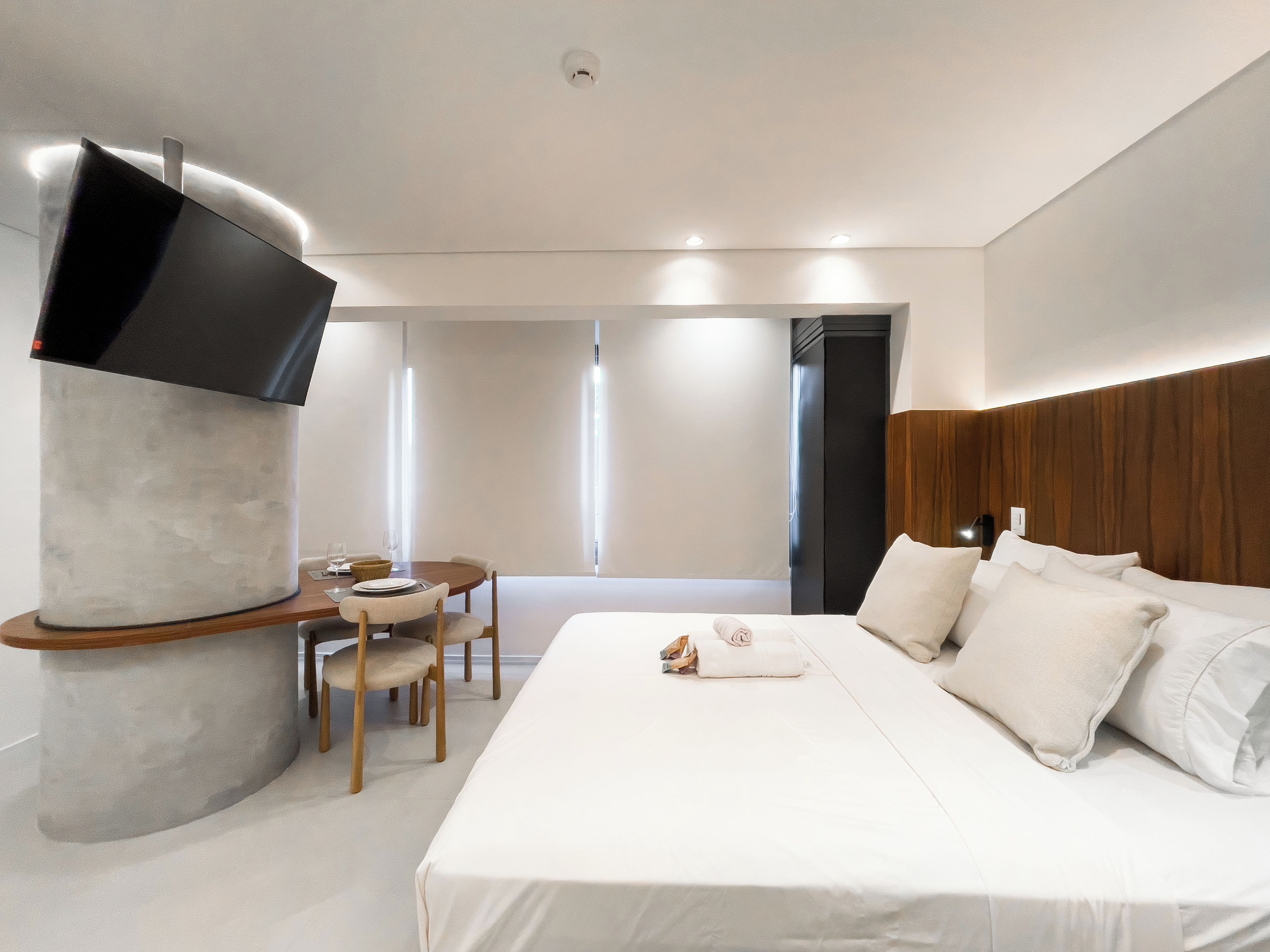 A modern hotel room with a large bed, a dining table, and a TV mounted on a cylindrical structure.