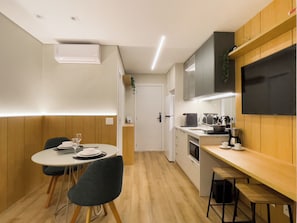 Cozinha compacta com área de refeições, painéis de madeira, forno embutido e televisão de tela plana.
