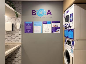 Uma área de lavanderia automática com uma placa afixada na parede exibindo o logotipo e o nome 'BioA', juntamente com diversas instruções e orientações de uso.