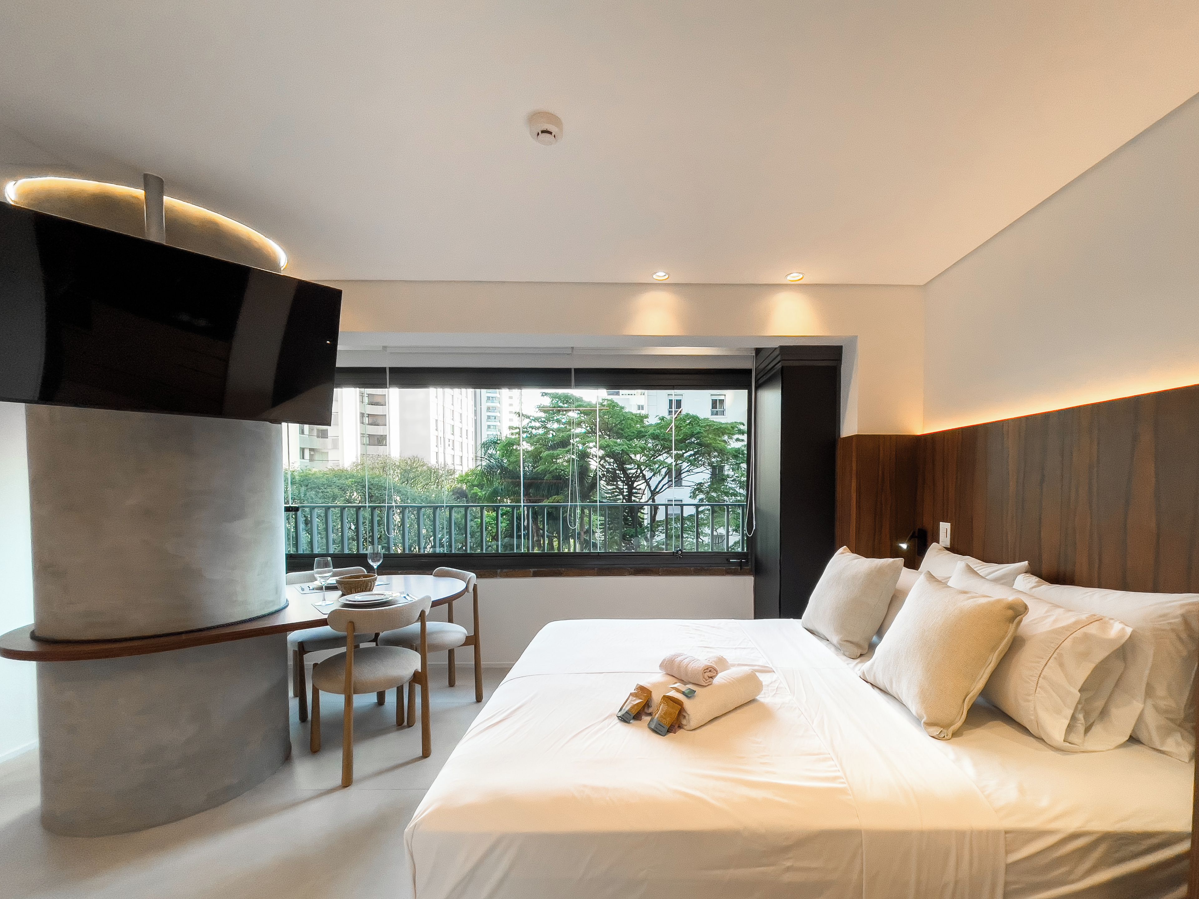 A modern hotel room with a large bed, a TV mounted on a pillar, a balcony with a view of buildings and trees, and a small table with chairs.