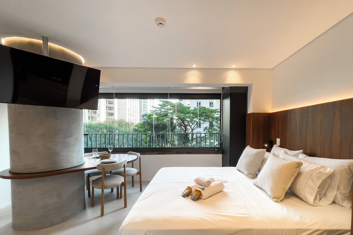 Una camera d'hotel moderna con un letto grande, una TV fissata a un pilastro, un balcone con vista su edifici e alberi, e un tavolino con sedie.
