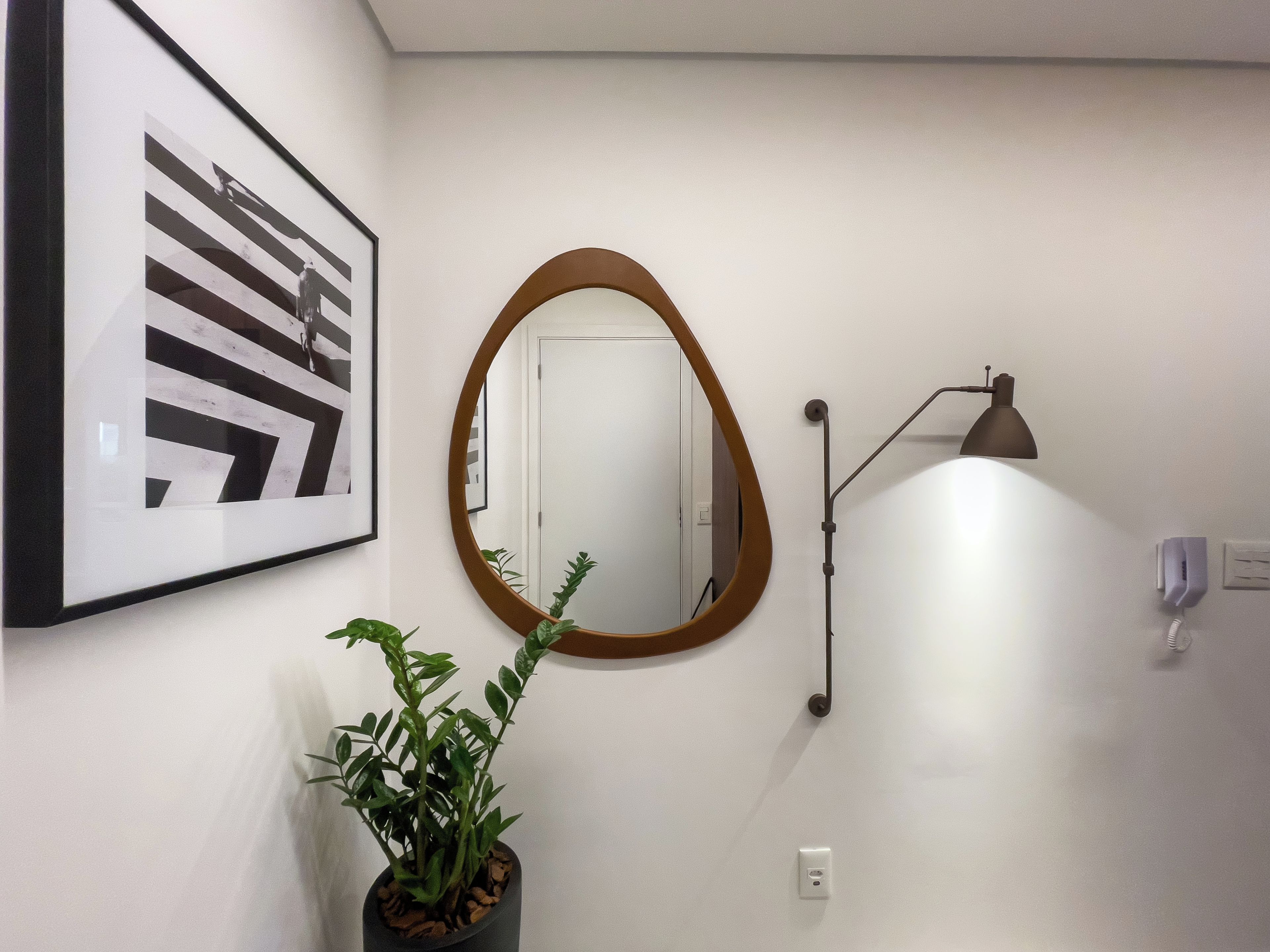 Um corredor com uma obra de arte abstrata emoldurada, um espelho redondo, um vaso de planta, uma luminária de parede e uma porta.