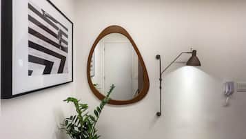 Um corredor com uma obra de arte abstrata emoldurada, um espelho redondo, um vaso de planta, uma luminária de parede e uma porta.