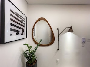 Un pasillo con una obra de arte abstracta enmarcada, un espejo redondo, una planta en maceta, una lámpara de pared y una puerta.